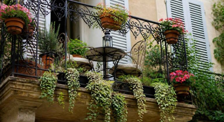 Balkon z ozdobną kutą balustradą, doniczkami kwiatów i zwisającą zielenią przy oknach z okiennicami